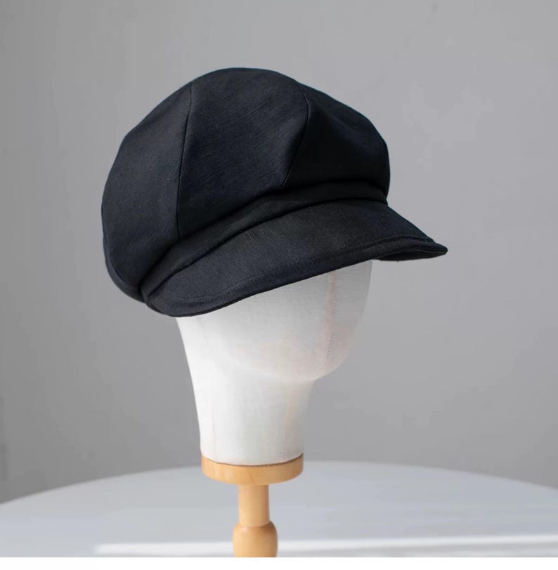 Puede incluir: Una gorra de newsboy de tela negra con una visera ligeramente curvada. La gorra est&aacute; en una cabeza de maniqu&iacute; blanca.