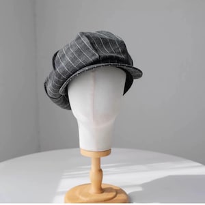 May include: A gray and white striped newsboy cap on a white mannequin head. The mannequin is on a wooden stand.
