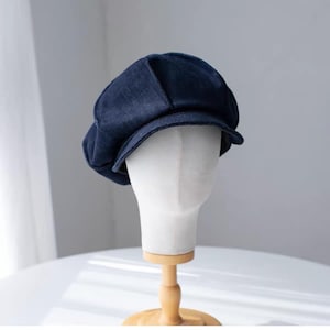 Puede incluir: Una gorra de estilo boina en tela vaquera azul oscuro con una corona estructurada de varios paneles y una visera corta y curva. El gorro presenta una forma redondeada clásica con costuras visibles a lo largo de los paneles.