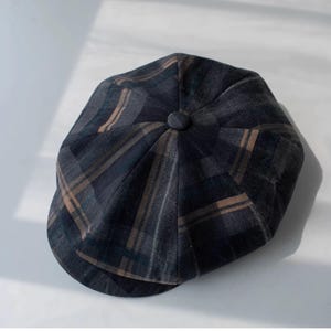 May include: A dark gray newsboy cap with a plaid pattern in shades of brown and blue. The cap has a rounded crown with a button on top and a short, curved brim. The fabric appears to be a textured material.