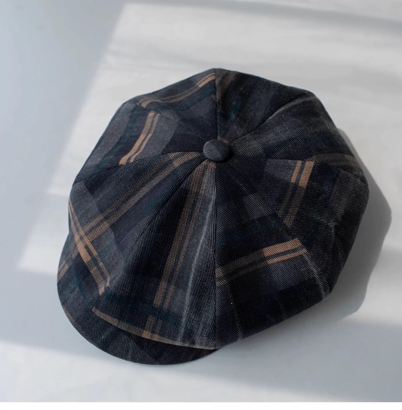 May include: A dark gray newsboy cap with a plaid pattern in shades of brown and blue. The cap has a rounded crown with a button on top and a short, curved brim. The fabric appears to be a textured material.