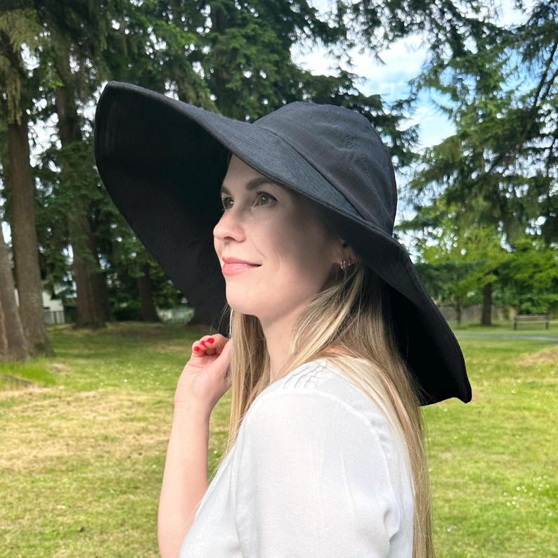 May include: A black sun hat with a wide brim. The hat is made of a soft material and is worn by a person outdoors. The background includes trees and grass.