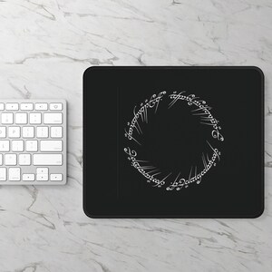 Op de afbeelding: Zwart rechthoekig muismatje met een wit cirkelvormig ontwerp met gestileerde tekst. Het ontwerp is een ring van tekst, waarschijnlijk uit een fantasy roman of film. Een wit toetsenbord is zichtbaar.