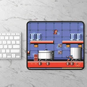 Op de afbeelding: Een blauwe en witte gepixelde videogame scène met een personage dat over een pot op een fornuis springt. Het personage draagt een rode hoed en een blauwe overall. De scène speelt zich af in een keuken met een blauwe betegelde vloer en witte kasten.