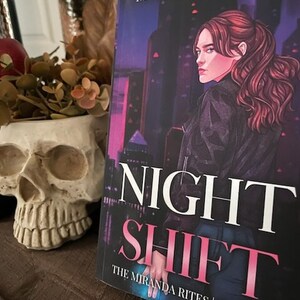 May include: A book cover with a dark purple background and a woman with long red hair looking to the left. The title of the book is "Night Shift" in white and pink letters. The text "The Miranda Rites | Book One" is in white letters on a black background.