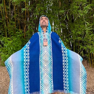 May include: A hooded poncho featuring a gradient of blue and teal tones with a central white geometric diamond pattern and vertical borders. The garment has a fringed hem, a buttoned neckline, and a rectangular cardboard tag hanging from the collar.