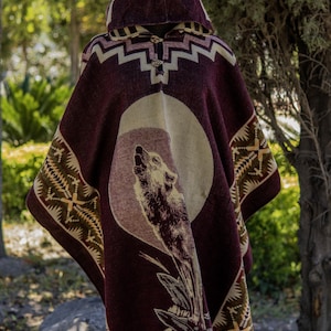 May include: A hooded poncho in shades of burgundy, beige, and brown, featuring a howling wolf design within a circular motif. The poncho has geometric patterns along the hood and edges, with a fringed hemline. The garment is displayed outdoors.