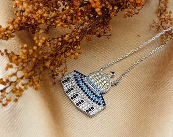 May include: A silver necklace with a pendant shaped like a dome with blue and white stones. The pendant is on a white fabric background.