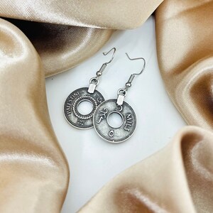 May include: Silver earrings with a coin design featuring the text "Palestine" and "5 Mils". The earrings are on a light brown fabric background.