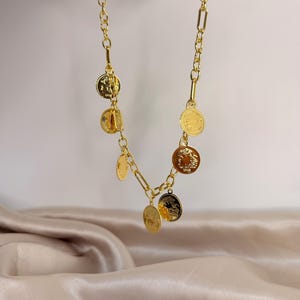 May include: Gold-toned necklace featuring coin pendants. The necklace has a chain link design with rectangular and oval links. The coin pendants are circular and have detailed engravings. The necklace is displayed against a neutral background.