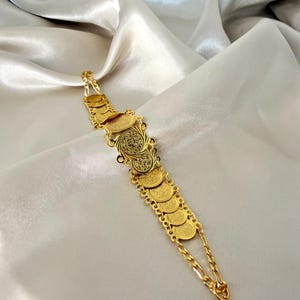 May include: A gold-colored bracelet featuring a series of coin-shaped charms and delicate chain links. The bracelet has an ornate design with intricate detailing on the coins. The jewelry is displayed on a satin-like fabric.