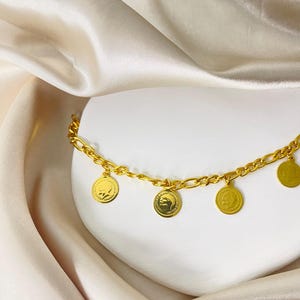 May include: A gold-colored anklet with a chain design and several dangling coin charms. The coins feature embossed designs. The anklet is displayed on a white surface with a cream-colored satin fabric in the background.