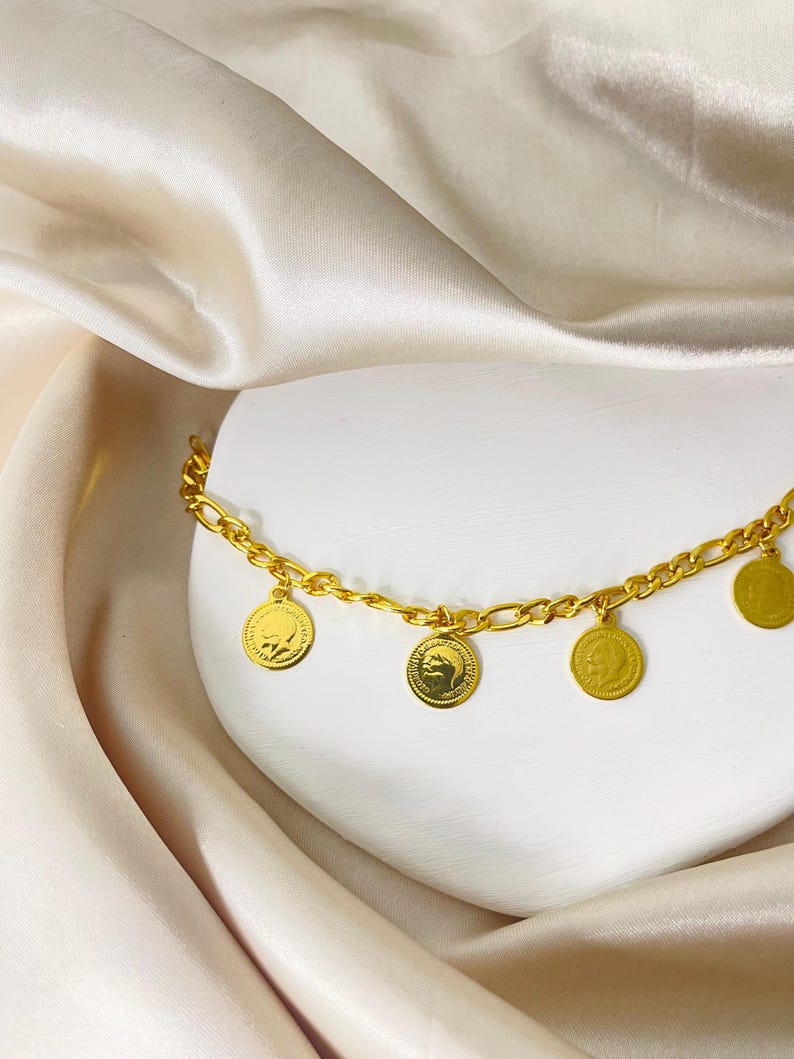 May include: A gold-colored anklet with a chain design and several dangling coin charms. The coins feature embossed designs. The anklet is displayed on a white surface with a cream-colored satin fabric in the background.