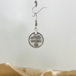 May include: Silver tone dangle earrings featuring a round coin with the word "Palestine" and the year "1927" in English. The coin also has Arabic text.