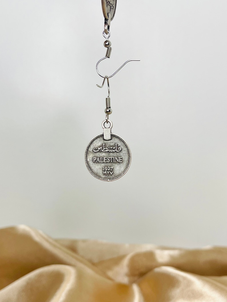 May include: Silver tone dangle earrings featuring a round coin with the word "Palestine" and the year "1927" in English. The coin also has Arabic text.