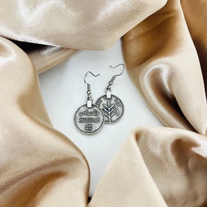 May include: Silver tone earrings featuring a round coin charm with the word "Palestine" and Arabic text on one side and a stylized olive branch on the other side.