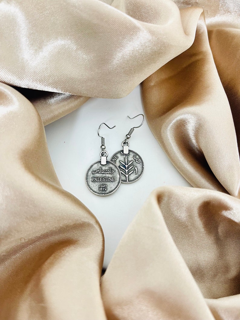 May include: Silver tone earrings featuring a round coin charm with the word "Palestine" and Arabic text on one side and a stylized olive branch on the other side.