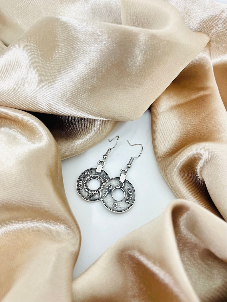 May include: Silver earrings with a coin design featuring the text "Palestine" and "5 Mils". The earrings are on a light brown fabric background.