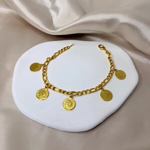 May include: A gold-colored bracelet with a chain design and several dangling coin charms. The coins feature a profile image. The bracelet is displayed on a white, textured surface against a neutral background.