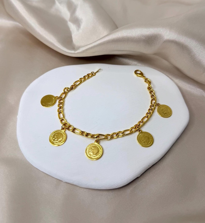 May include: A gold-colored bracelet with a chain design and several dangling coin charms. The coins feature a profile image. The bracelet is displayed on a white, textured surface against a neutral background.