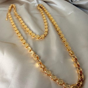 May include: A gold-colored necklace with a repeating pattern of circular and oval links. The necklace is displayed on a satin surface, and the links have a textured design. The chain is a long style, suitable for layering or wearing alone.