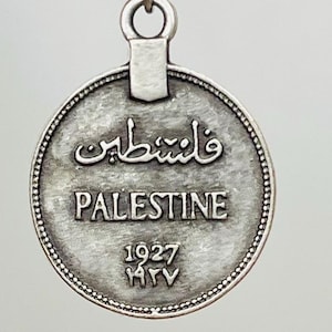 May include: Silver coin with a loop at the top. The coin has the word "Palestine" and the year "1927" inscribed on it. The coin also has Arabic text on it.