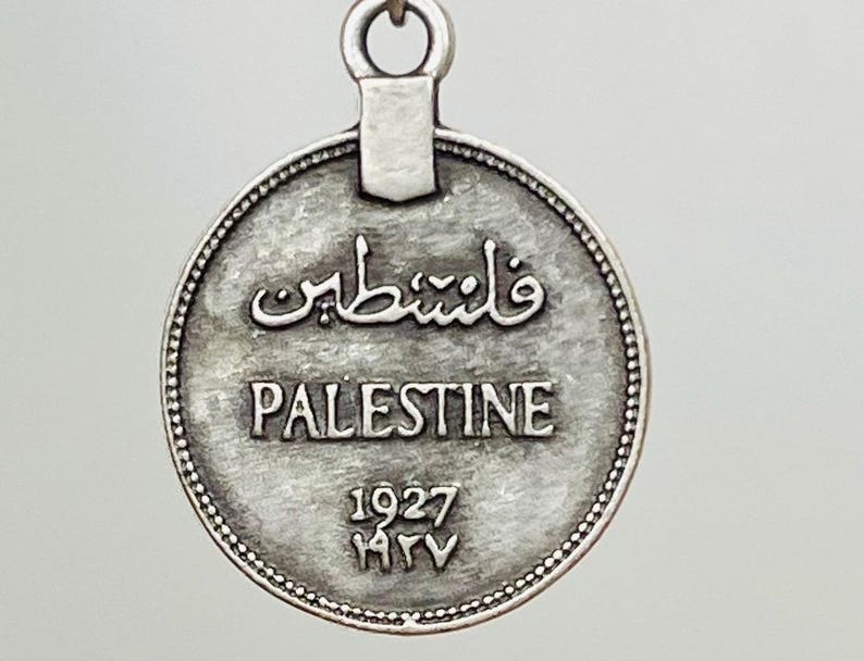 May include: Silver coin with a loop at the top. The coin has the word "Palestine" and the year "1927" inscribed on it. The coin also has Arabic text on it.