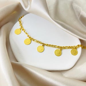 May include: A gold-colored bracelet with a chain design and five circular charms. The charms have a detailed design. The bracelet is displayed on a white, curved surface against a backdrop of draped, off-white fabric.