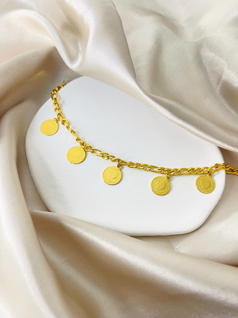 May include: A gold-colored bracelet with a chain design and five circular charms. The charms have a detailed design. The bracelet is displayed on a white, curved surface against a backdrop of draped, off-white fabric.