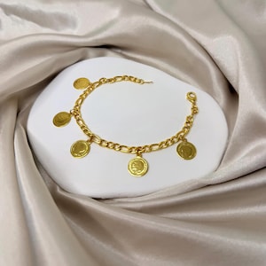 May include: Gold-toned bracelet featuring a chain design with multiple circular coin charms. The bracelet is displayed on a white surface against a backdrop of a silky, beige fabric. The coin charms have a detailed design.