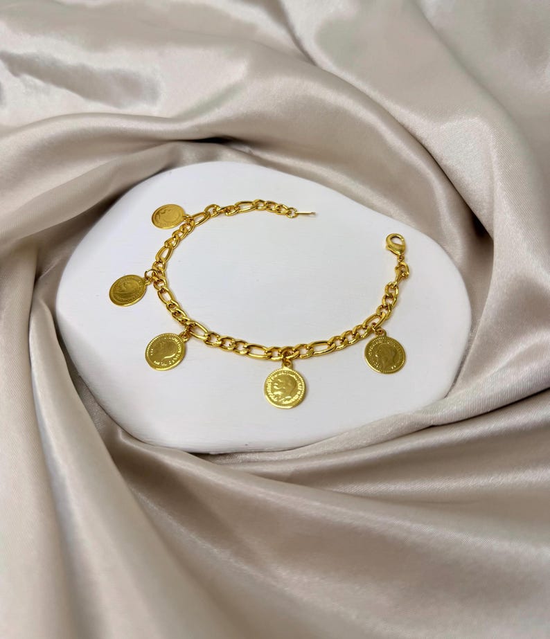 May include: Gold-toned bracelet featuring a chain design with multiple circular coin charms. The bracelet is displayed on a white surface against a backdrop of a silky, beige fabric. The coin charms have a detailed design.