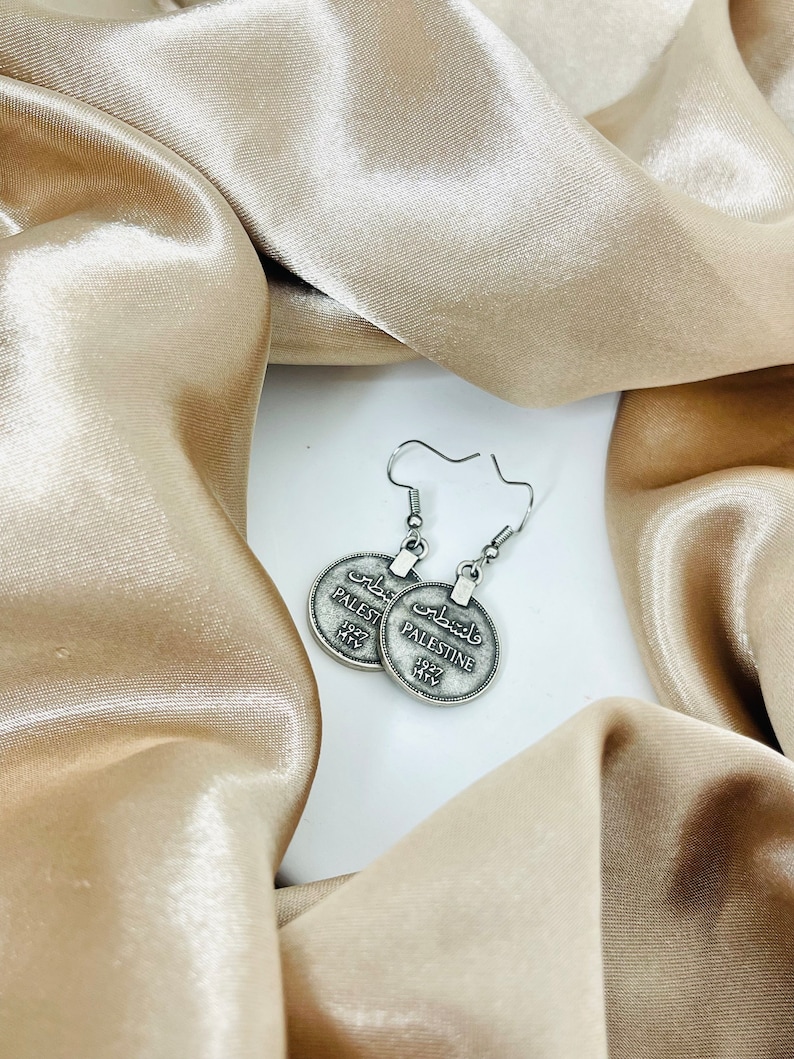May include: Silver earrings with a round coin design featuring the word "Palestine" in both English and Arabic. The coins are attached to silver hooks.