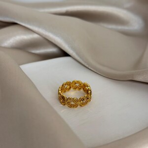 May include: A gold-colored ring with a repeating floral design. The ring is set against a white surface and a beige satin background. The ring's intricate details are highlighted by the lighting.