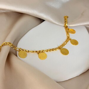May include: A gold-colored bracelet with a chain design and several small, round, coin-like charms. The bracelet is displayed on a white surface with a beige satin fabric in the background. The charms have a detailed design.