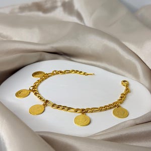 May include: A gold-colored charm bracelet featuring a chain with alternating oval and rectangular links. The bracelet has several round, gold-colored charms with a detailed design. The bracelet is displayed on a white surface with a satin fabric background.