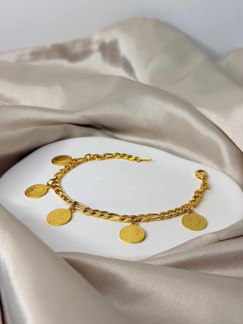 May include: A gold-colored charm bracelet featuring a chain with alternating oval and rectangular links. The bracelet has several round, gold-colored charms with a detailed design. The bracelet is displayed on a white surface with a satin fabric background.