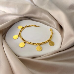 May include: Gold-toned bracelet with a curb chain design and several circular charms. The charms appear to have a detailed design. The bracelet is displayed on a white surface against a backdrop of draped, silky fabric.
