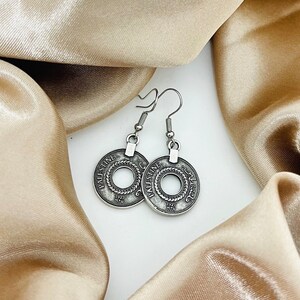 May include: A pair of silver earrings with a circular design featuring the text "Palestine" and Arabic script. The earrings are hanging from a white surface.