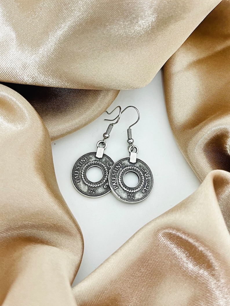 May include: A pair of silver earrings with a circular design featuring the text "Palestine" and Arabic script. The earrings are hanging from a white surface.