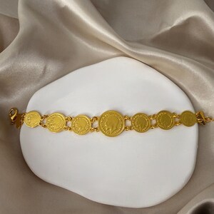May include: A gold-colored coin bracelet featuring multiple circular coins with embossed profiles. The bracelet is displayed on a white, curved surface against a backdrop of a silky, beige fabric. The bracelet is a jewelry item.