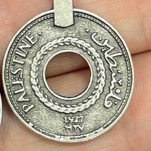 May include: A silver-toned pendant charm featuring a round coin with a hole in the centre. The coin has the word "PALESTINE" and Arabic text around the edge. The year "1927" is inscribed on the bottom.