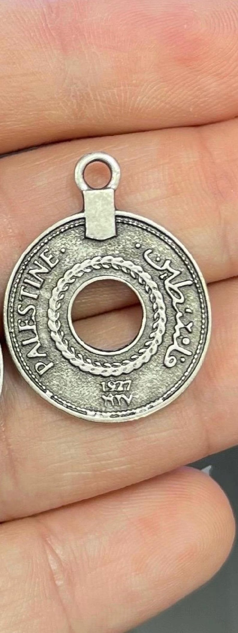 May include: A silver-toned pendant charm featuring a round coin with a hole in the centre. The coin has the word "PALESTINE" and Arabic text around the edge. The year "1927" is inscribed on the bottom.
