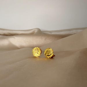 May include: A pair of gold-colored stud earrings. Each earring features a circular design with a detailed relief of a profile and a spiral border. The earrings are displayed on a beige, textured surface.