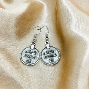 May include: Silver tone earrings with a round coin design. The coins feature the word "Palestine" in English and Arabic, along with the year "1927".