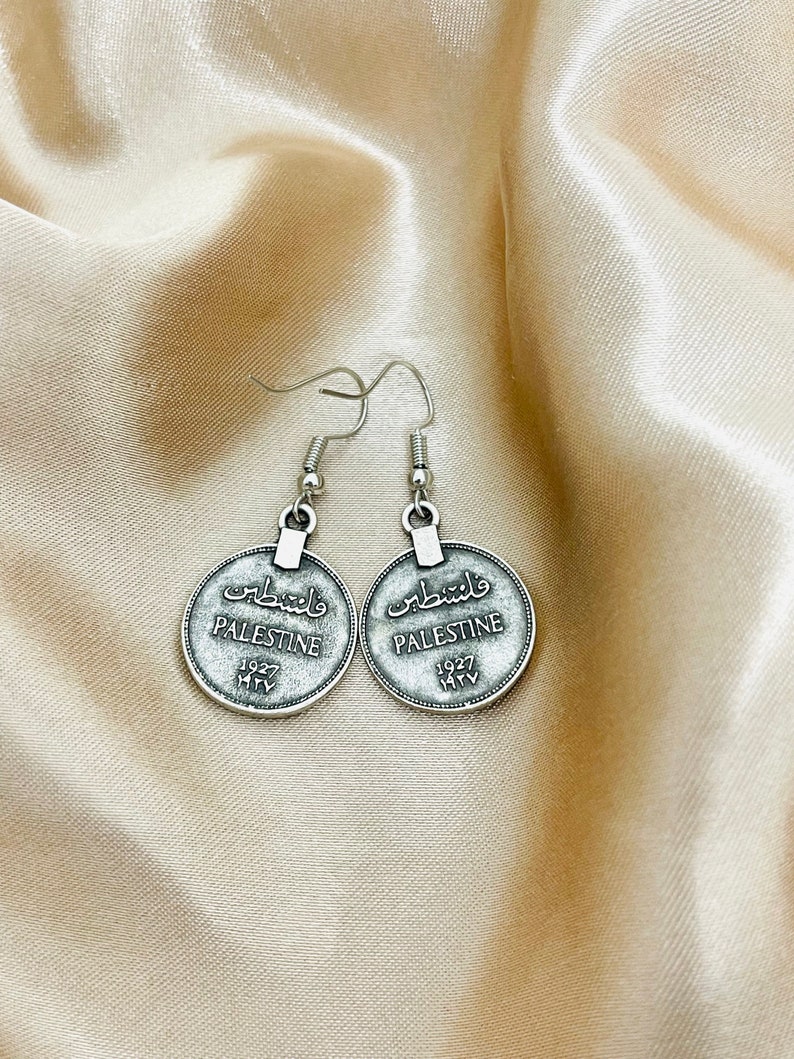 May include: Silver tone earrings with a round coin design. The coins feature the word "Palestine" in English and Arabic, along with the year "1927".