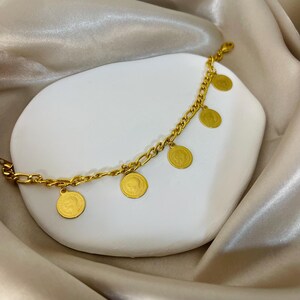 May include: A gold-colored bracelet with a chain link design and several dangling coin charms. The coins have a detailed embossed design. The bracelet is displayed on a white, organic-shaped surface against a backdrop of draped, satin fabric.