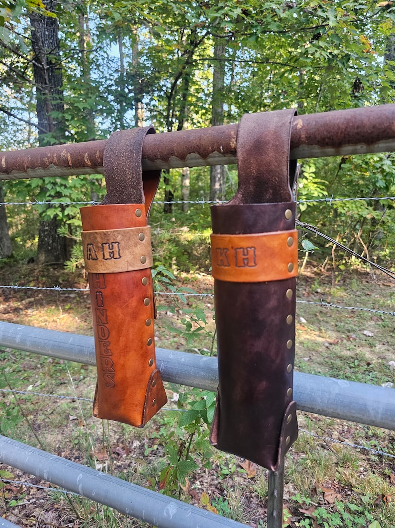 Pu&ograve; includere: Due porta bottiglie in pelle marrone con rivetti metallici e cinghie appese a una recinzione metallica arrugginita. I porta bottiglie sono personalizzati con le iniziali "AH" e "KH" e la parola "HUNDRED" &egrave; impressa sul porta bottiglie di sinistra.