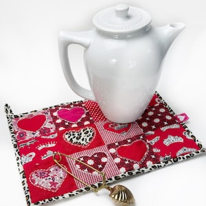 May include: A white teapot sits on a red and white quilted hot pad with a heart pattern. A gold tea infuser is on the table next to the hot pad.