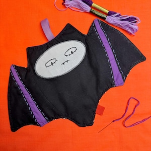 May include: A black bat-shaped fabric item with purple detailing and a stitched face, on an orange background. Purple embroidery thread and a needle are present, indicating a craft project. A small red tag is attached.