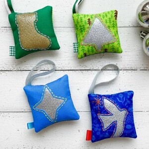 Peut inclure: Ornements de Noël colorés. Les décorations sont des coussins carrés avec des motifs festifs : une chaussette, un arbre, une étoile et une colombe. Des paillettes argentées mettent en valeur les motifs. Deux boules argentées sont également présentes.